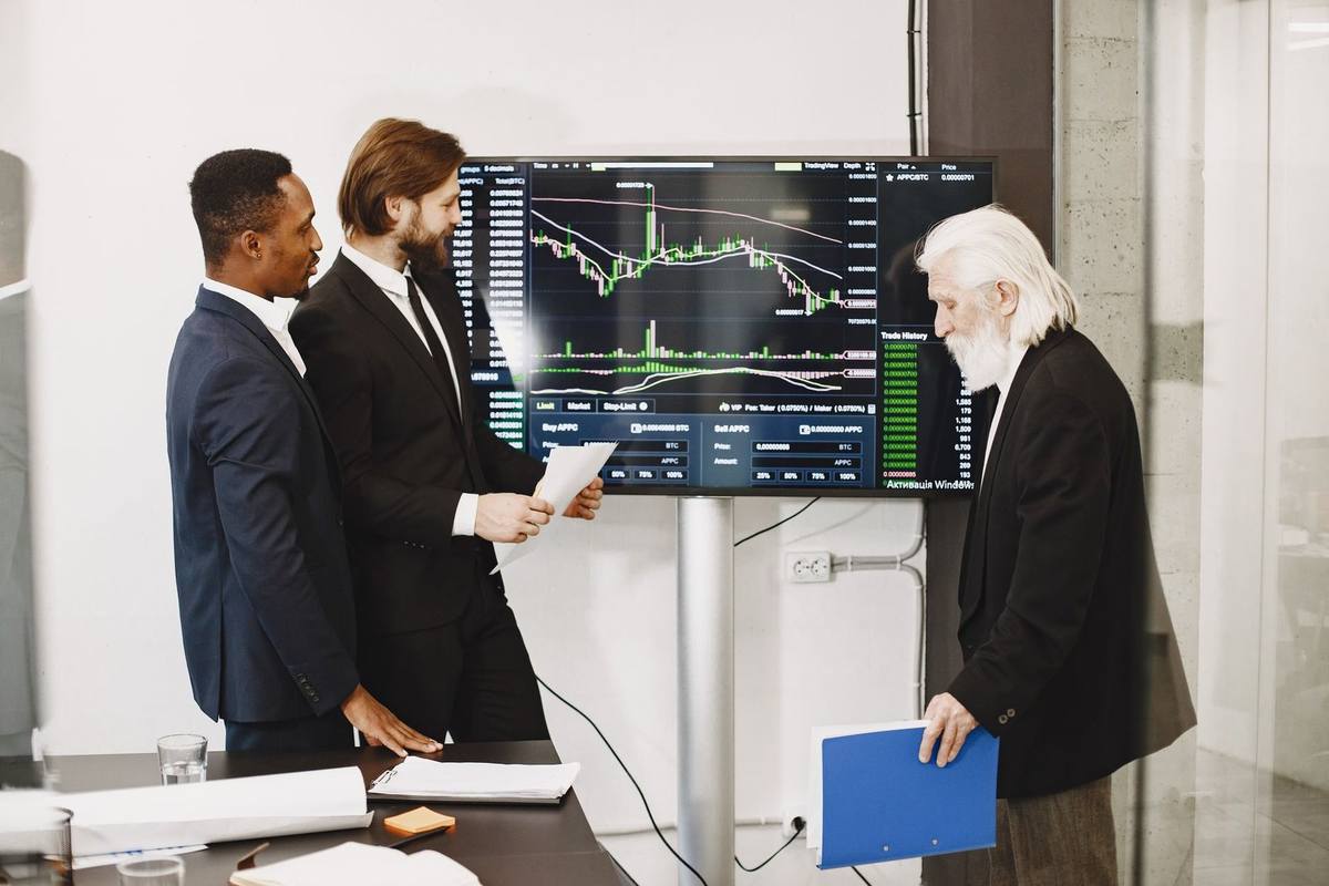 Trois hommes en réunion devant un écran de graphiques financiers, documents à la main, dans un bureau moderne Trois hommes en réunion devant un écran de graphiques financiers, documents à la main, dans un bureau moderne