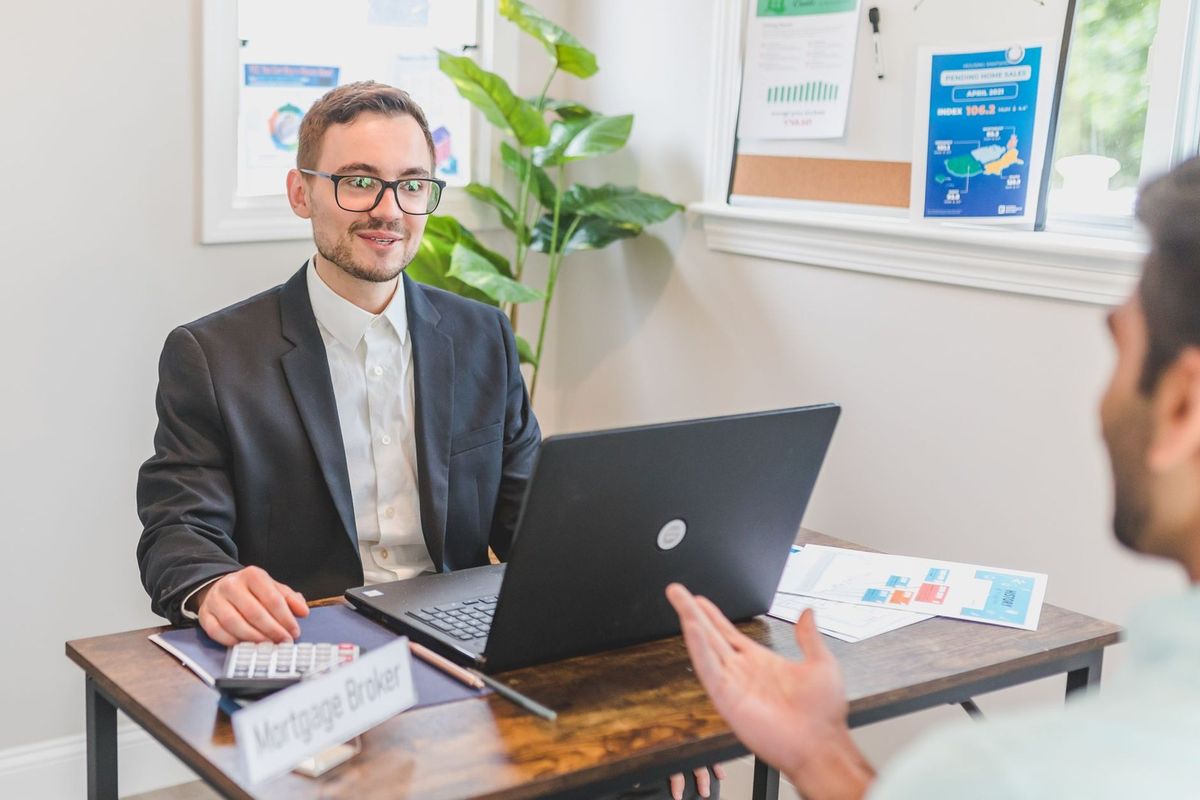 Homme en costume devant ordinateur portable, échange avec un autre homme, documents et plante sur la table Homme en costume devant ordinateur portable, échange avec un autre homme, documents et plante sur la table