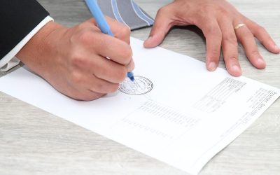 Main droite écrivant sur un document avec un stylo bleu, main gauche posée dessus, table en bois