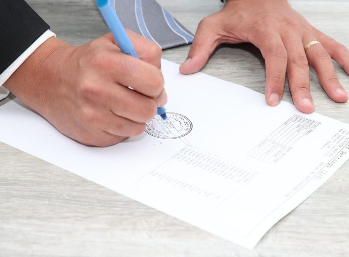 Main droite écrivant sur un document avec un stylo bleu, main gauche posée dessus, table en bois